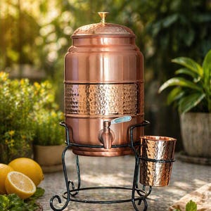 May include: A copper water dispenser with a hammered texture, a spigot, and a lid, set on a black wrought iron stand. A matching copper cup sits in a holder. Lemons and greenery are in the background.