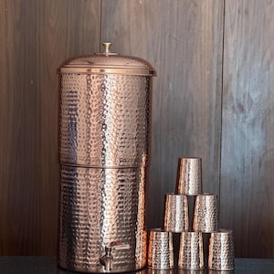 May include: A copper water dispenser with a hammered texture, a spigot, and a lid. The dispenser is accompanied by a stack of eight matching copper cups. The copper finish has a warm, reddish-brown hue, and the items are arranged on a dark surface.