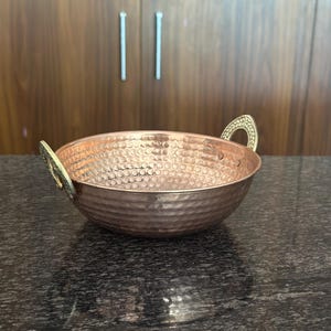 Könnte beinhalten: Eine gehämmerten Kupferschale mit zwei Messinggriffen. Die Schale hat eine strukturierte Oberfläche und eine runde Form. Die Kupferfarbe kontrastiert mit den Messinggriffen. Die Schale wird wahrscheinlich zum Servieren von Speisen oder als Dekorationsartikel verwendet.