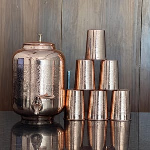 May include: A copper water dispenser with a spigot and lid, next to a pyramid of six matching copper cups. The dispenser and cups have a hammered texture. The items are on a dark surface with a wooden background.