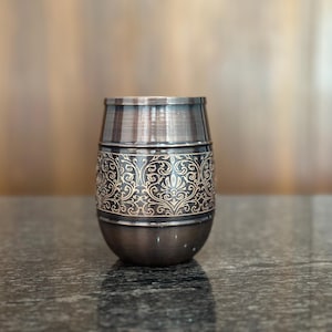 May include: A decorative metal cup with an ornate, swirling gold pattern on a dark background. The cup has a bronze finish and a rounded base. The cup is likely used for drinking.