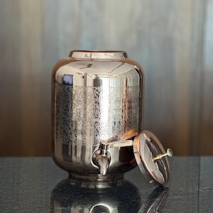May include: A polished silver beverage dispenser with an ornate floral pattern. The dispenser has a spigot, a removable lid, and a reflective surface. The container is sitting on a dark, glossy surface.