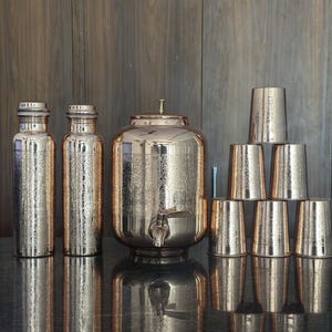 May include: A collection of copper-colored drinkware, including two tall bottles, a large water dispenser with a spigot, and a stack of seven drinking glasses. The items have a textured surface and are arranged on a dark surface.