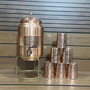 May include: A copper water dispenser with a spigot and a set of six matching copper cups. The dispenser sits on a gold-colored stand. The cups are stacked in a pyramid shape. The dispenser and cups have a hammered texture.