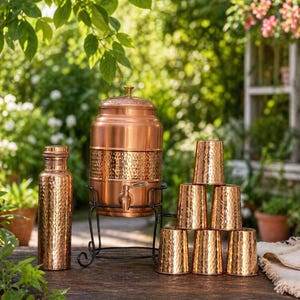 May include: A copper water dispenser with a spigot, a matching water bottle, and a stack of copper cups. The dispenser sits on a black metal stand. The items have a hammered texture and are set on a wooden table.
