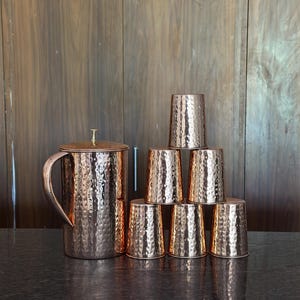 May include: A copper pitcher with a lid and handle, and a set of seven matching copper cups. The pitcher and cups have a hammered texture. The set is displayed on a dark surface against a wooden background.