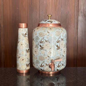 Könnte beinhalten: Eine zylindrische Wasserflasche und ein großer Wasserspender mit Zapfhahn, beide mit hellblauem Hintergrund, weiß-braunem Blumenmuster, kupferfarbenen Metallsockeln und einem passenden Deckel mit einem kleinen messingfarbenen Knopf.