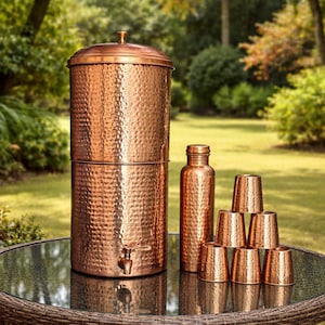 May include: A copper water filter with a hammered texture, a matching bottle, and six small copper cups are arranged on a glass-topped table. The water filter has a spigot and a lid. The items are outdoors in a garden setting.