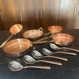 Puede incluir: Una colección de utensilios de cocina y cucharas de servir de cobre. El juego incluye una sartén grande, sartenes pequeñas, un cucharón y cucharas. Las piezas de cobre tienen una textura martillada y asas de latón. Las cucharas tienen un cuenco plateado pulido y mangos de cobre.