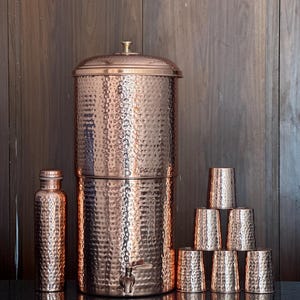 Könnte beinhalten: Ein Kupfer-Wasserfiltersystem mit gehämmertem Muster, eine passende Flasche und sechs Kupferbecher. Der Wasserfilter hat einen Wasserhahn und einen Deckel mit einem kleinen Griff. Die Artikel sind auf einer dunklen Oberfläche angeordnet.