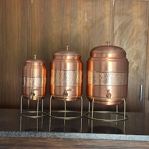 May include: Three copper beverage dispensers of different sizes. Each has a hammered band, a tap, and a lid with a small handle. They stand on gold-coloured metal stands, arranged in a row against a wooden cabinet backdrop.