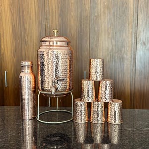 May include: A hammered copper water dispenser with a lid and spigot, resting on a metal stand, accompanied by a matching hammered copper bottle and six small hammered copper tumblers arranged in a pyramid formation on a dark surface.