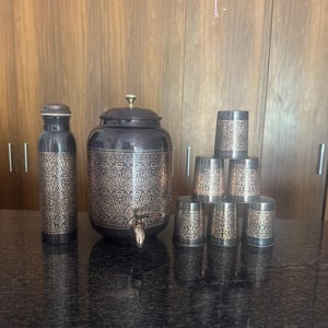 Könnte beinhalten: Ein schwarzes und goldenes Getränkeset mit einem Wasserspender, einer Flasche und sechs passenden Bechern. Das Set hat ein dekoratives Damastmuster. Der Spender hat einen Wasserhahn und einen Deckel mit einem goldenen Knopf. Die Becher sind gestapelt.