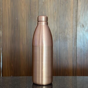 May include: A copper-colored water bottle with a rounded body and a screw-on cap. The bottle has a smooth, polished finish and stands upright on a dark surface. The background is a wooden wall.