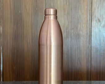May include: A copper-colored water bottle with a rounded body and a screw-on cap. The bottle has a smooth, polished finish and stands upright on a dark surface. The background is a wooden wall.