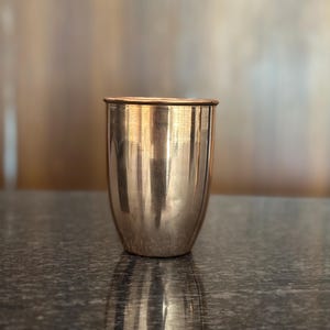 May include: A copper-colored, cylindrical cup with a slightly flared rim. The cup has a smooth, polished surface that reflects the light. The cup is sitting on a dark, textured surface.