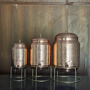 May include: Three copper-colored hammered metal beverage dispensers of varying sizes. Each has a spigot and sits on a gold-colored metal stand. The dispensers have domed lids and are displayed against a dark wood background.