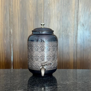 May include: A dark blue decorative water dispenser with a silver spigot and lid. The dispenser has an ornate, repeating pattern in a lighter color. The dispenser is sitting on a dark, speckled surface.