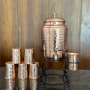 May include: Copper water dispenser set with six matching cups. The dispenser has a hammered texture and sits on a black metal stand. The cups are stacked in front of the dispenser. The set is displayed on a dark surface.