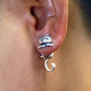 Puede incluir: Pendientes de plata con forma de gato que cuelgan con un pequeño dije de gato en la parte inferior.