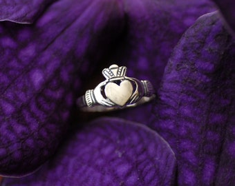 Puede incluir: Un anillo Claddagh de plata con un diseño de corazón, manos y corona. El anillo está sobre un pétalo de flor morado.