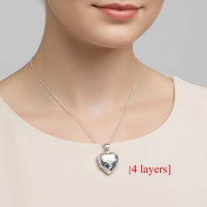 Puede incluir: Collar con colgante de corazón plateado y cadena fina. El relicario está cerrado y tiene un diseño detallado en la parte delantera. El texto "[4 layers]" es visible.