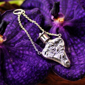 May include: A silver heart-shaped perfume bottle with an ornate floral design hangs from a silver chain. The bottle is resting on a purple orchid flower.