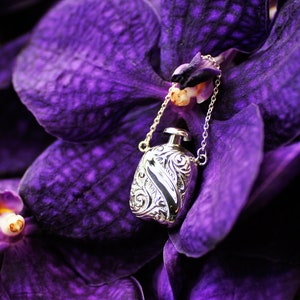 May include: A silver pendant in the shape of a bottle with an ornate design hangs from a silver chain. The pendant is resting on a purple orchid flower.