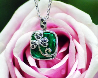 Puede incluir: Un collar de plata con un colgante de trébol verde y negro. El colgante está engastado en un marco cuadrado y tiene un diseño detallado. El collar está fotografiado sobre una rosa rosa.