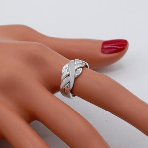 K&ouml;nnte beinhalten: Ein polierter Ring in Silberoptik mit einem mehrteiligen Puzzle-Design, bestehend aus ineinandergreifenden, geflochtenen Segmenten, die sich um den Finger legen. Die Metalloberfl&auml;che ist glatt und reflektierend und bildet ein komplexes Muster.