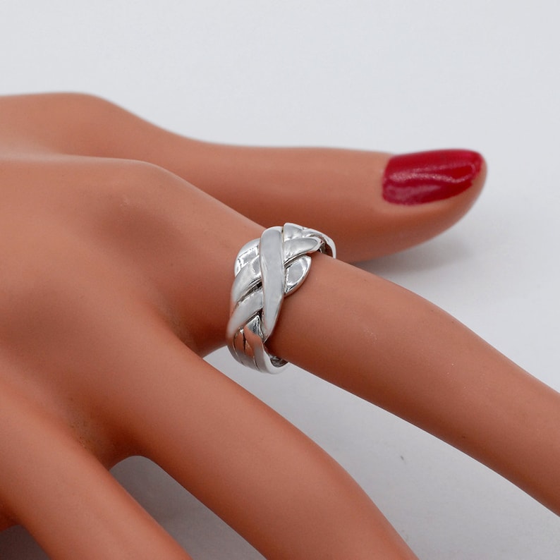 K&ouml;nnte beinhalten: Ein polierter Ring in Silberoptik mit einem mehrteiligen Puzzle-Design, bestehend aus ineinandergreifenden, geflochtenen Segmenten, die sich um den Finger legen. Die Metalloberfl&auml;che ist glatt und reflektierend und bildet ein komplexes Muster.