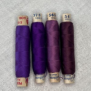 Puede incluir: Cuatro carretes de hilo morado, que varían en tonalidad de claro a oscuro. Cada carrete tiene una etiqueta de papel con números y texto, incluyendo "Gütermann Ideal-S". Los carretes están dispuestos verticalmente sobre una superficie texturizada gris claro.
