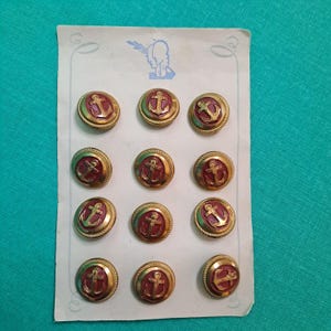 Puede incluir: Un conjunto de doce botones de latón vintage con un diseño de ancla. Cada botón presenta un centro rojo con un ancla dorada y un borde dorado en forma de cuerda. Los botones están dispuestos en una tarjeta blanca con una ilustración azul.