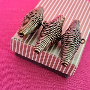 Pode incluir: Três contas de metal douradas com um design tecido. As contas estão em uma pequena caixa com um padrão listrado.