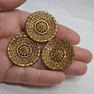 May include: Three gold-toned metal buttons with intricate, circular designs. Each button features a central, raised, starburst pattern surrounded by a ring of smaller, raised circles.