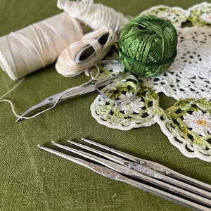 Op de afbeelding: Een set van vijf zilveren haaknaalden met de merknaam "Boye" erop gedrukt. De naalden liggen op een groene stoffen ondergrond naast een witte gehaakte doily, een bol groene garen en een spoel wit garen.