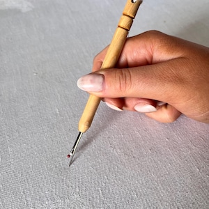 May include: A close-up of a hand holding a wooden threader with a red tip. The threader is being used to thread a needle.