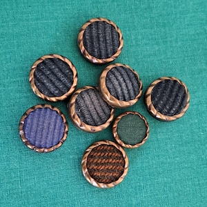 Op de afbeelding: Assortiment van acht ronde, met stof beklede knopen met een koperkleurige metalen rand. De knopen hebben verschillende patronen en kleuren, waaronder bruin, groen, blauw en grijs.