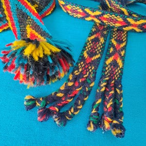 May include: Close-up of a colorful woven textile with a blue background. The textile features a pattern of yellow, red, and green diamonds on a dark blue base. A fluffy pom-pom with yellow, blue, and red strands is also visible.