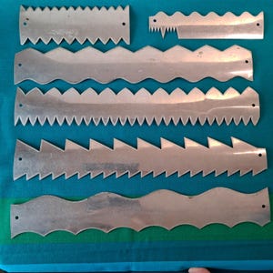 Puede incluir: Colección de siete cuchillas de acero inoxidable con bordes dentados y ondulados. Las cuchillas están dispuestas sobre una superficie a rayas azul y turquesa. Cada cuchilla presenta un diseño diferente, incluyendo zigzags, triángulos y ondas redondeadas.