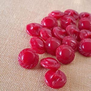 May include: A pile of glossy, red buttons with a textured, circular design. The buttons are scattered on a tan, woven fabric. These vintage-style buttons are ideal for sewing and craft projects.