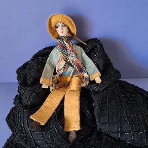 Op de afbeelding: Een vintage pop gekleed in een lichtblauwe jas, bruine broek en een kleurrijke sjaal. De pop draagt een gele hoed en heeft een geschilderd gezicht. Het zit op een zwarte, getextureerde stof met een blauwe achtergrond.