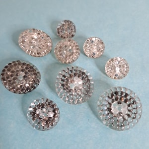 May include: A selection of clear, round buttons with a textured, bubble-like design. The buttons are of varying sizes and are arranged on a light blue surface. The buttons have a reflective quality, catching the light.