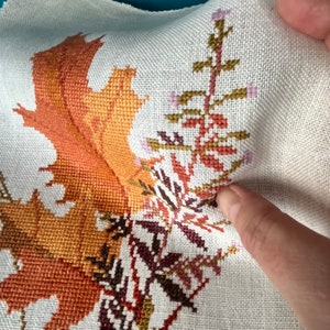 May include: Close-up of a cross-stitch embroidery featuring a detailed design of autumn leaves in shades of orange, brown, and gold, with a smaller design of green and brown leaves in the foreground.