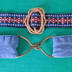 May include: A woven belt with a tan oval buckle and a blue and black patterned band. The belt also features a light blue fabric and a gold-coloured metal clasp. The belt is displayed on a green surface.