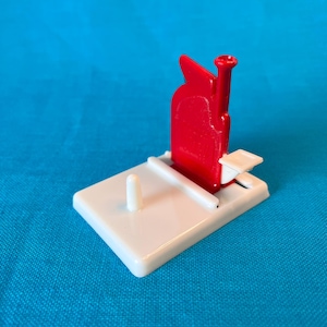 May include: A white plastic sewing thread holder with a red plastic thread guide and a small white plastic post. The thread guide has a small hole at the top for threading.