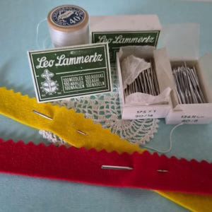 May include: A collection of sewing supplies including a spool of white thread, two boxes of Leo Lammertz needles labeled 175 x 1 90/14 and 134 R 80/8, and two strips of felt fabric with scalloped edges in bright yellow and red with needles inserted.
