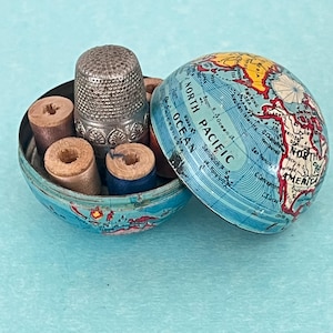 Op de afbeelding: Een metalen wereldbolhouder in twee helften, met een kaart van de Noordelijke Stille Oceaan en Noord-Amerika. De binnenkant bevat een zilverkleurige metalen vingerhoed met een getextureerd oppervlak en diverse kleine houten klossen naaigaren.