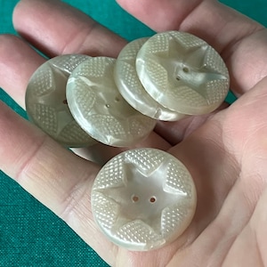 May include: A collection of five pearlescent, star-shaped buttons. Each button features a raised star design in the center, surrounded by a textured pattern. The buttons are round with two holes for sewing. The buttons are held in a hand.