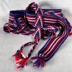 2 handgeweven linten. Vikingversieringen van wol. Riemen in Sami-stijl. Zweeds. Jaren 70. Gestreept en geruit patroon. Blauw, rood en wit. Kwastjes.